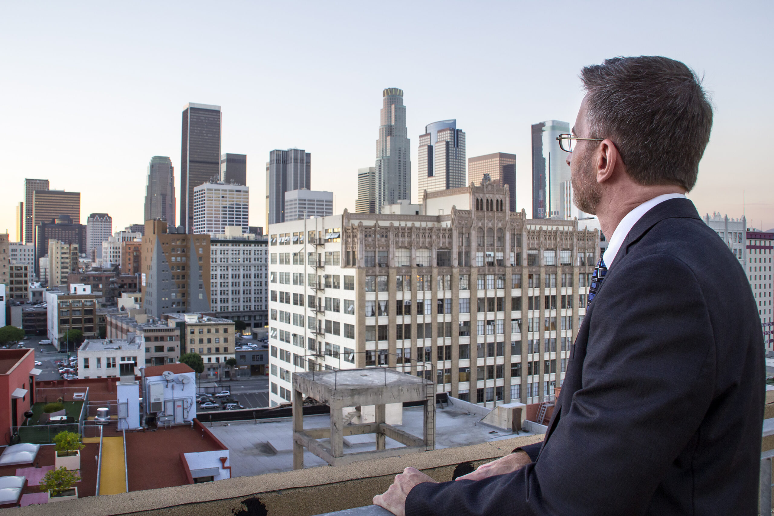Businessman or architect looking at real estate gentrification zone of Downtown Los Angeles.  The man is dressed in a suit.  The view is during blue hour after sunset at the historic core of the city.  The man looks like he is a city developer or a politician. The business logos have been removed from the buildings.