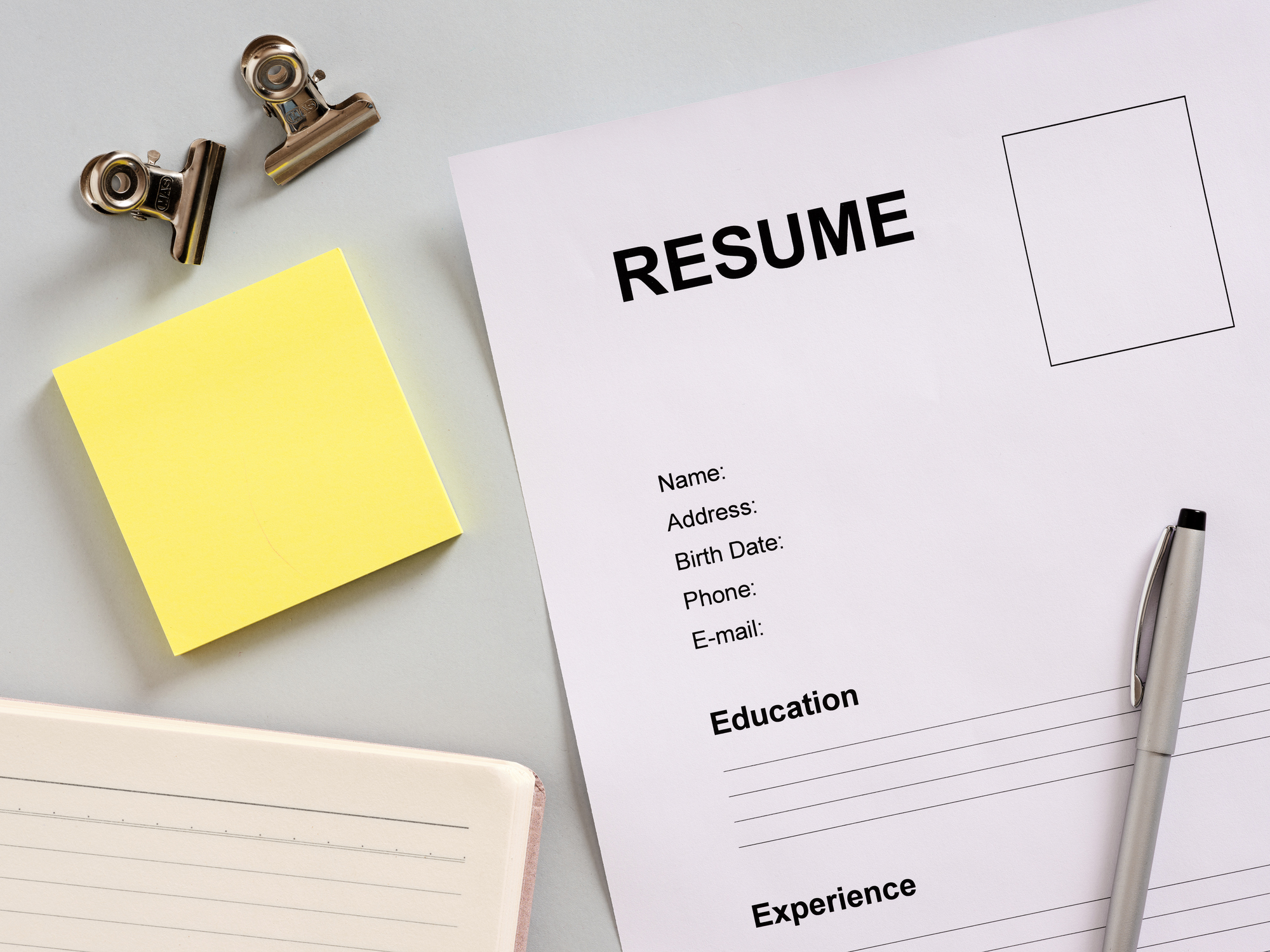 Resume, pen, notebook, and sticky notes arranged on a gray office desk, awaiting details to be filled in for job applications